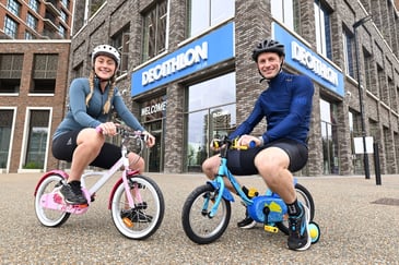 A woman and man sit on two small bicycles wearing white helmets, in front of a Decathlon shop sign that is blue and white